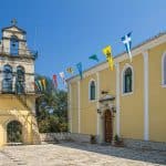 Traditional Greek Orthodox church in Corfu with a stone bell tower, colorful flags, and a bright blue sky in the background.